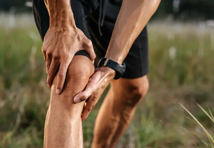 Un corridore si tiene il ginocchio, dolorante, non riesco a piegare il ginocchio. Mani sudate, orologio sportivo, campo erboso.
