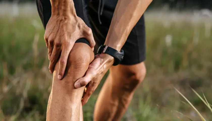 Un corridore si tiene il ginocchio, dolorante, non riesco a piegare il ginocchio. Mani sudate, orologio sportivo, campo erboso.