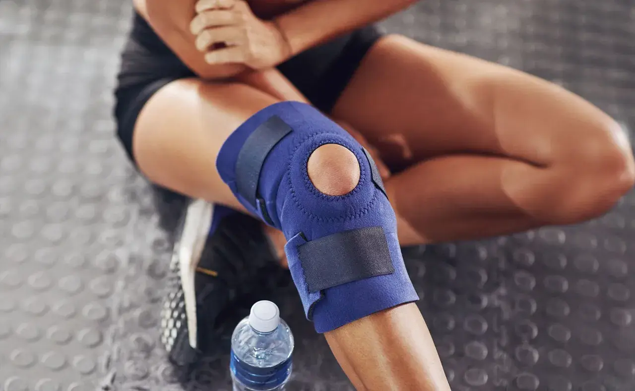 Donna con tutore al ginocchio blu, seduta su pavimento gommato. Considerare le controindicazioni del tutore ginocchio prima dell'uso.