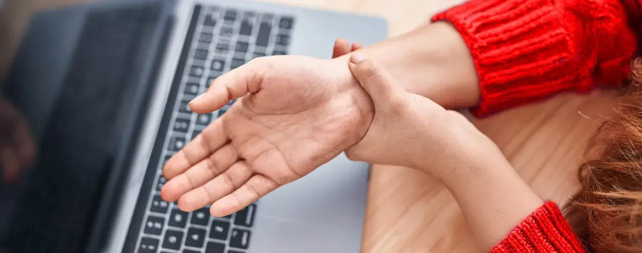 Mano che si tiene il polso, forse per dolori o fitte al polso, vicino a un laptop.