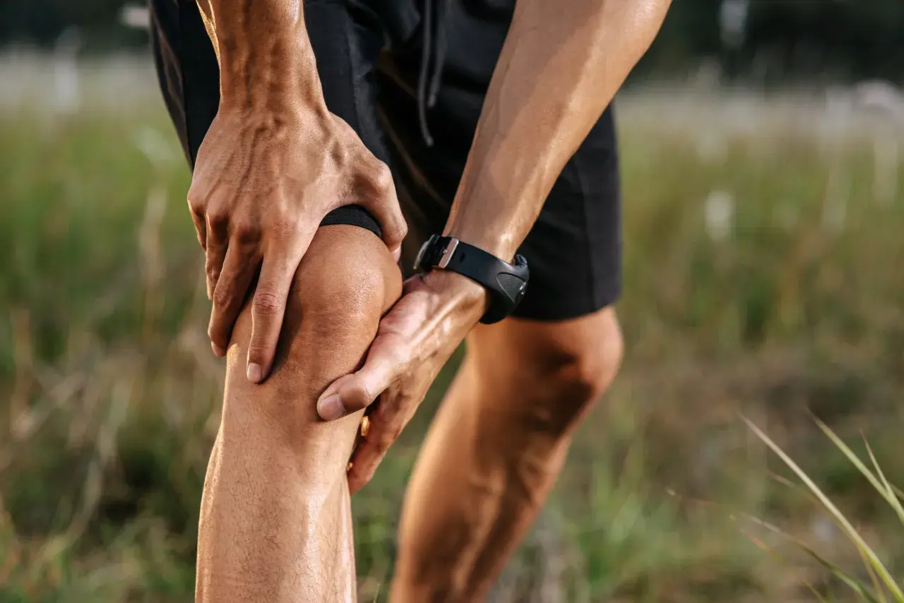 Un corridore si tiene il ginocchio, dolorante, non riesco a piegare il ginocchio. Mani sudate, orologio sportivo, campo erboso.